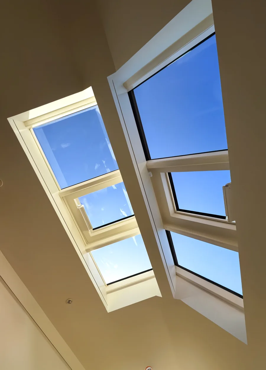 Looking up at a vaulted ceiling with two white-framed rooflights set in deep linings, their upper sashes open on motorised chain actuators right at the apex