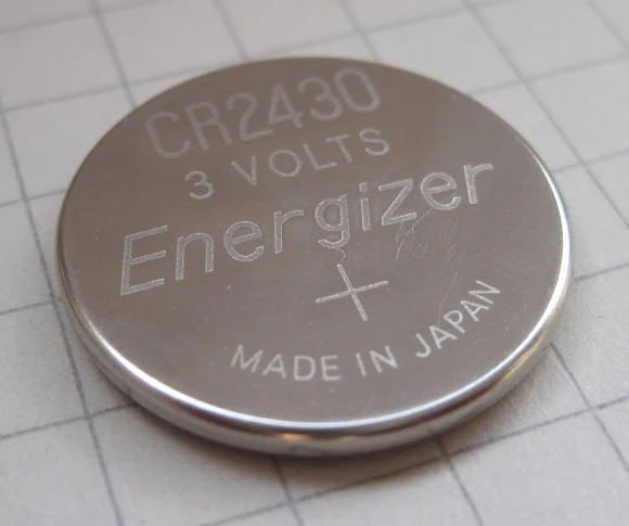 A CR2430 lithium coin cell battery photographed on a 7mm grid showing its 24mm diameter.