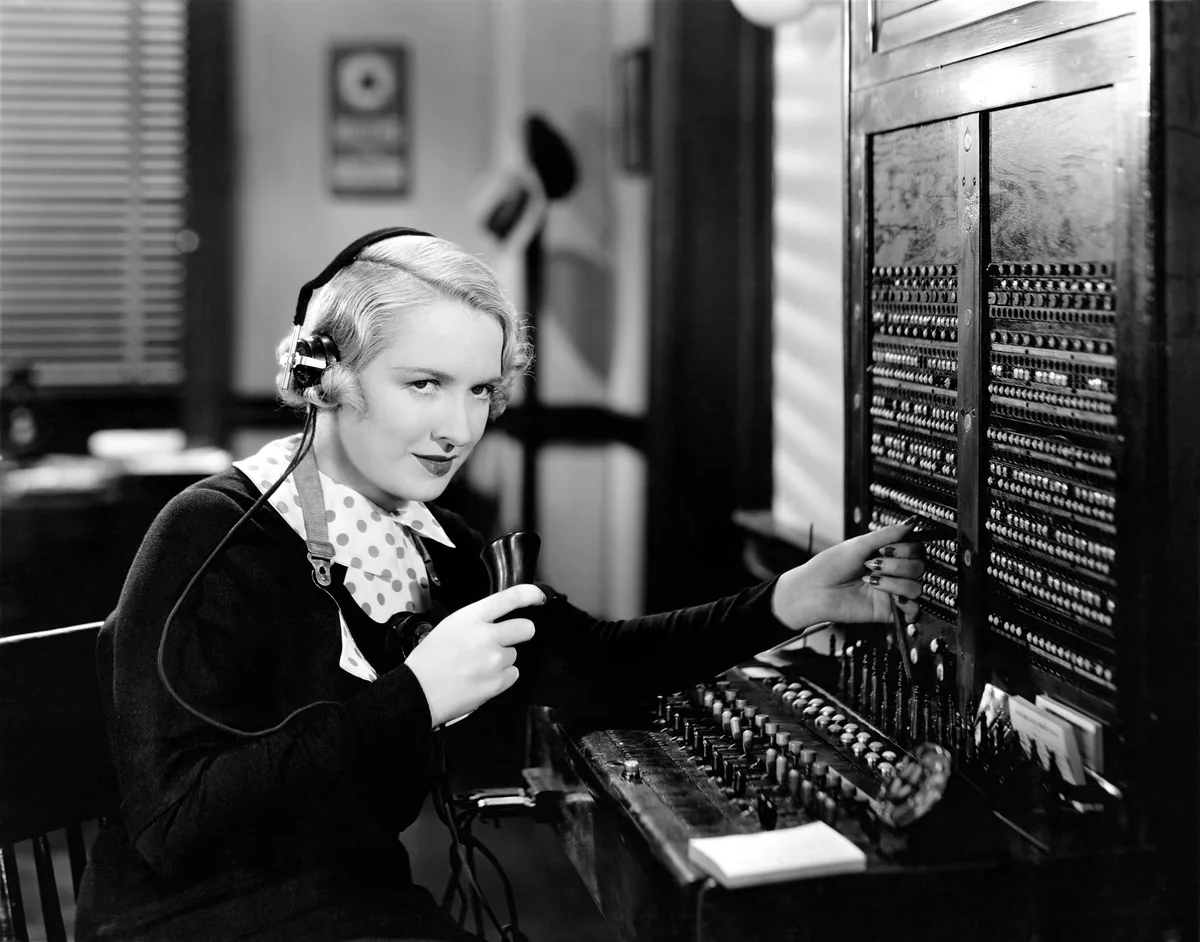 A telephone operator at an old-fashioned switchboard, in black and white