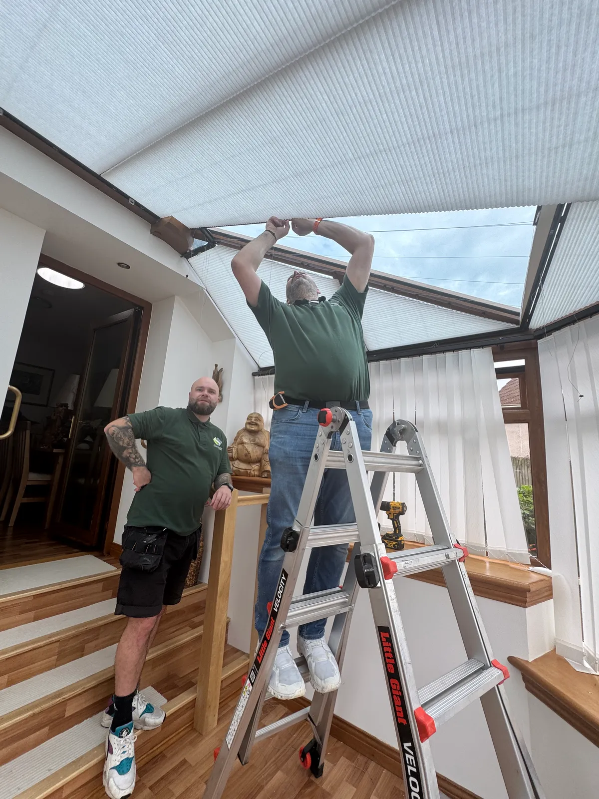 Two Scottish Shutter Company installers fitting cellular roof blinds to a conservatory roof in Perthshire, one working from a ladder with the shaped blinds already in place above